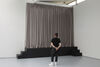 A photograph of the corner of gallery two. We can see two sets of small set of black steps leading to a black platform. At the top of the platform is a beige hanging curtain, as though a stage. A visitor stands looking at this installation, wearing black clothing and white trainers.