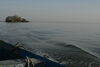 A film still, taken from a wooden boat, of a calm sea with a small island to the top left hand side on the horizon line, which is about a third from the top of the image. A small part of the boat can be seen on the bottom left. The sky is cloudless and the sea and sky are a similar grey-blue.