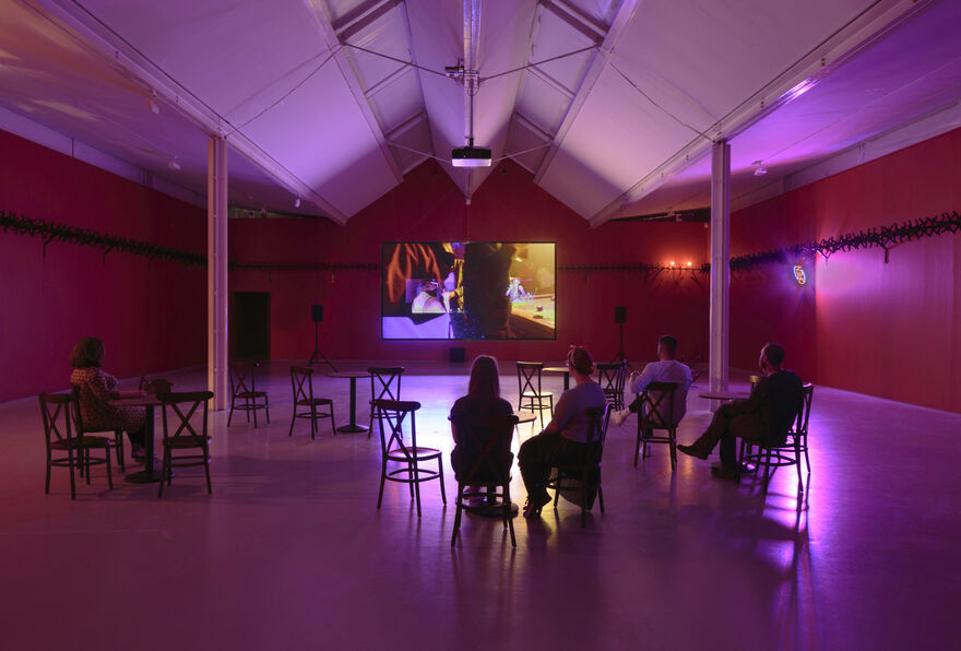 A wide shot photograph of gallery two, showing the gallery with red walls and cabaret-style seating with small tables in the floorspace. A few people sit and face a screen with a projected film.