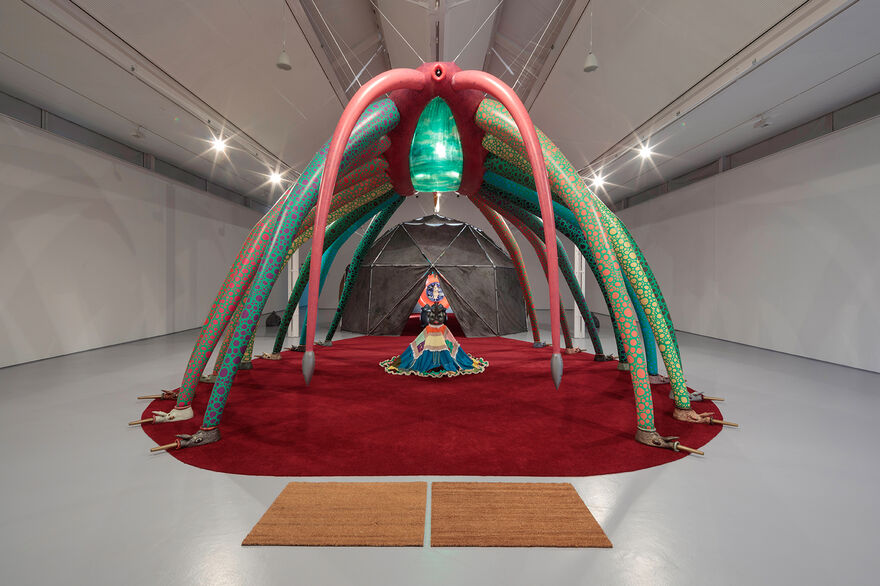 Photograph of gallery two, showing a large installation, in the centre of the space. On a big red carpet, a freestanding inflatable insect-like form towers over a geodesic dome containing video and ceramic elements.