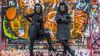 Two figures dressed in black, wearing gorilla masks, stand in front of a wall covered in graffiti. One has their arms folded and the other an arm on their hip.