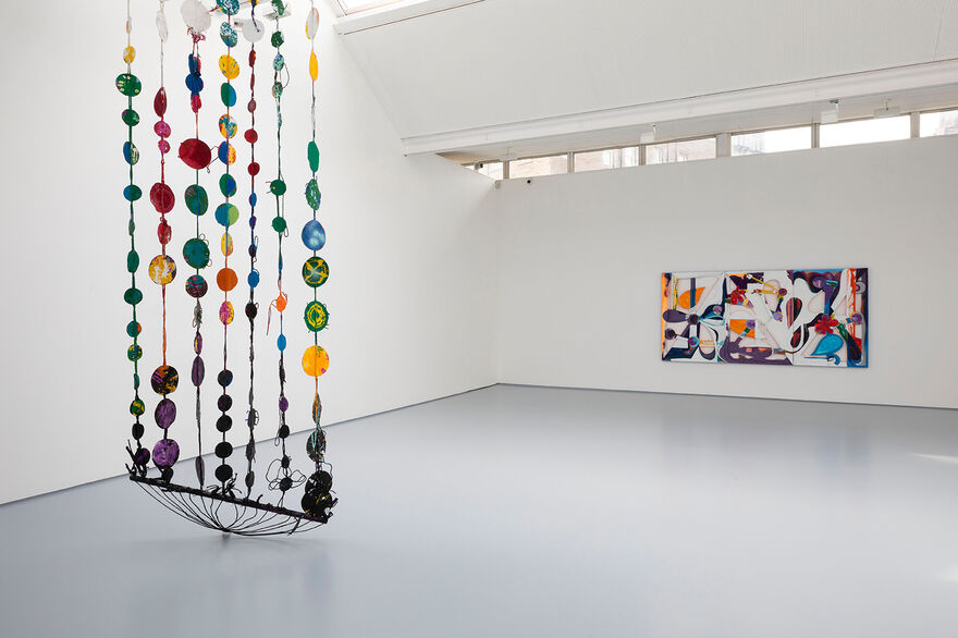 Installation documentation showing a hanging hammock form to the left hand side of the image, a colourful sculpture composed of painted found materials and circle motifs, braided together in strands. In the background to the left of the bright gallery, on a white wall, is a rectangular artwork mainly white, purple, orange and blue.