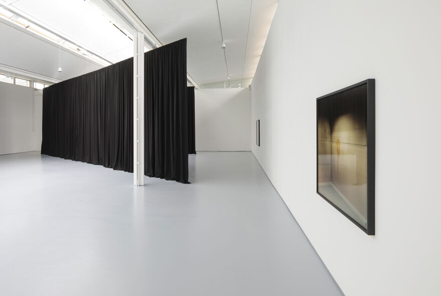 Photograph shows an installation in a brightly lit gallery with white walls and grey floor. There are large black pleated curtains hanging in the space, nearly to the ceiling. These are hung in broad sweeping diagonals across the large floorspace. On the wall to the right is a photograph framed.