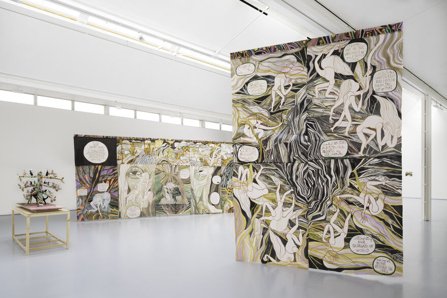 A hanging artwork on the right hand side of the image has watercolour figures and writing. To the left of the image can be seen another hanging work strung across the gallery. On a small wooden plinth to the left hand side of the image is a textile work.